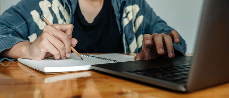 A student writing notes by hand at a desk with an AI chatbot open on a laptop nearby